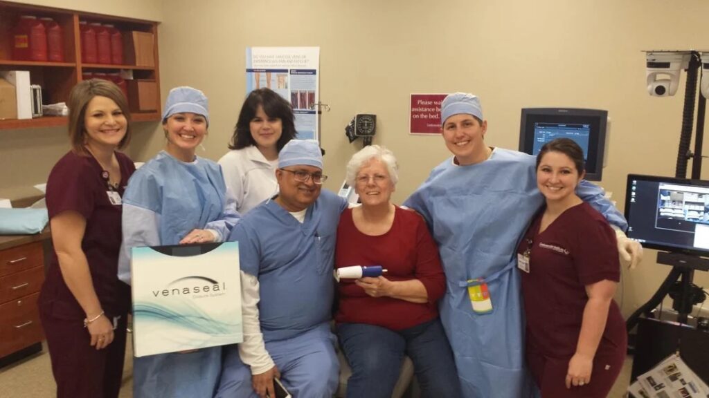 A group of smiling cardiologists in scrubs gathered around a seated patient in a clinical setting at the Cardiovascular Institute of the South, showcasing a sense of teamwork and heart health care.