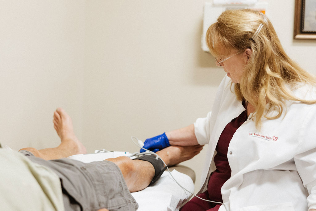 A healthcare professional is performing a medical procedure on a patient's leg in a cardiology clinical setting.