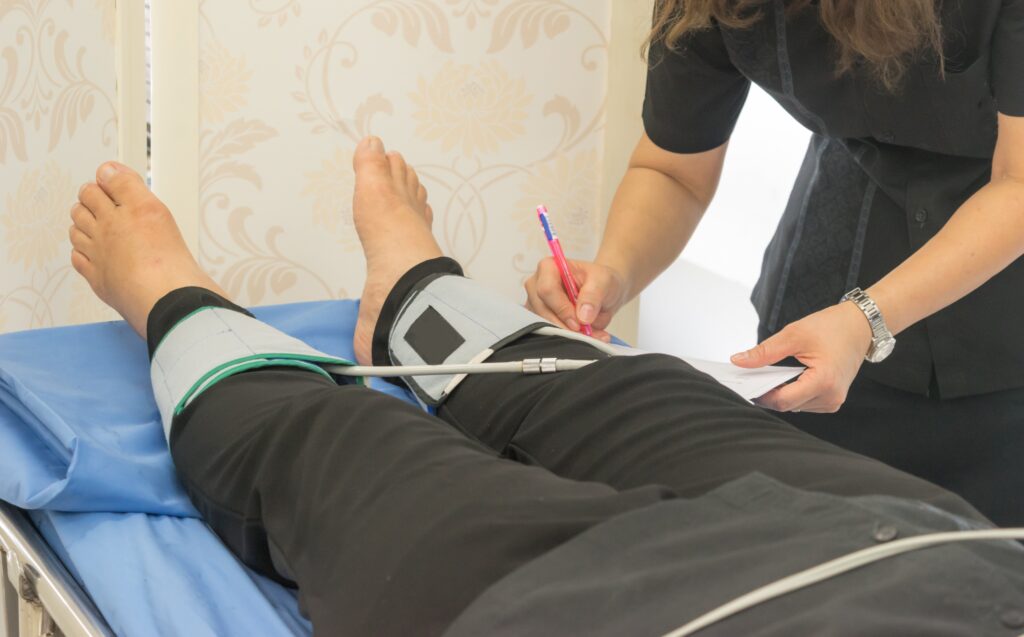 A patient lying down with a blood pressure cuff on their leg while a healthcare worker from the Cardiovascular Institute of the South takes notes during a medical examination for venous disease.