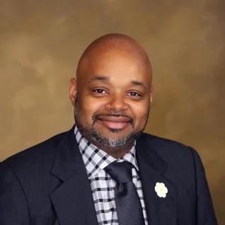 A professional portrait of a smiling man with a beard, dressed in a suit with a checkered tie and a flower on the lapel, specializing in Cardiology.