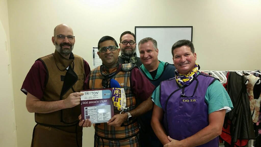 Group of five smiling individuals in colorful aprons and costume pieces, holding a cardiology event flyer and posing for a photo in a room with clothes racks in the background.