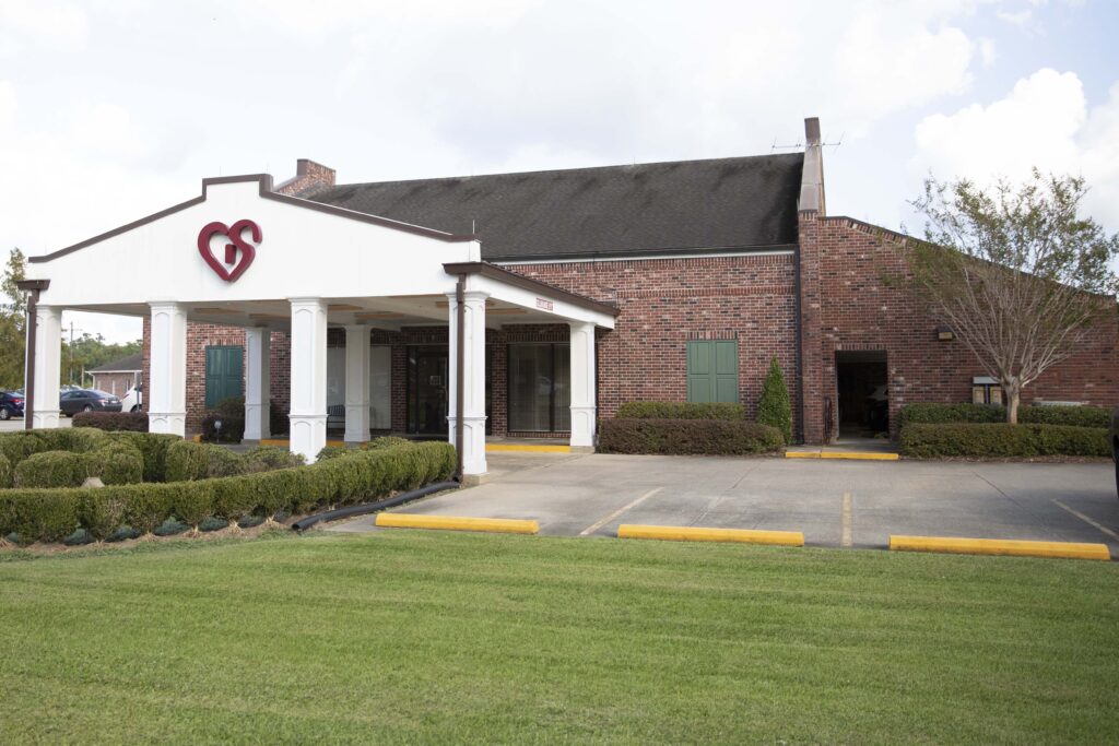 A single-story brick building with a white portico featuring a heart-shaped logo, serving as the entrance to a commercial establishment on a bright day with a partly cloudy sky.