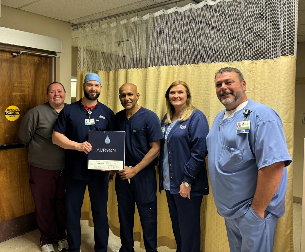 A group of five medical professionals, smiling, standing in a clinic room with a curtain in the background. one of them is holding an award plaque with the name "auryon" on it.