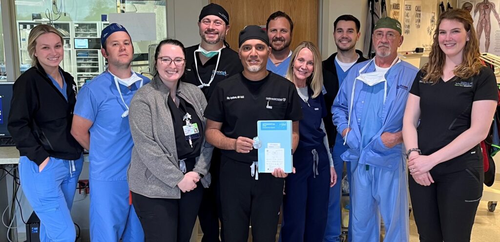 A group of nine medical professionals, including doctors and nurses specializing in heart failure CCM therapy, in scrubs, smiles for a photo in a hospital corridor; one holds a certificate.