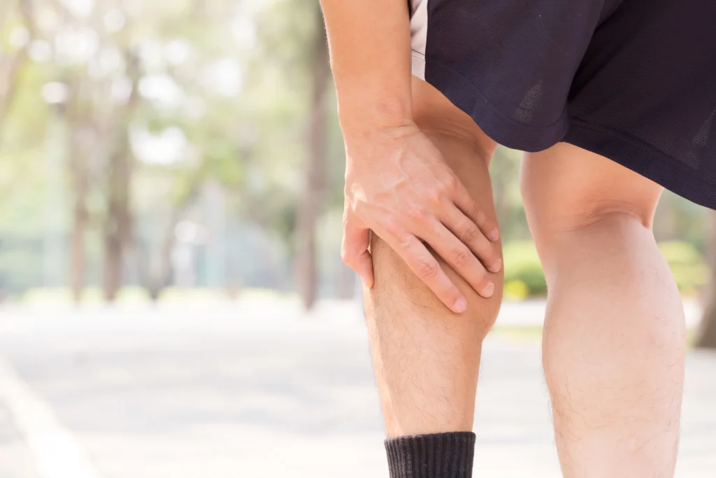 A person holding their hamstring in discomfort, possibly experiencing a muscle cramp or strain, outdoors in a park-like setting near the Cardiovascular Institute of the South.