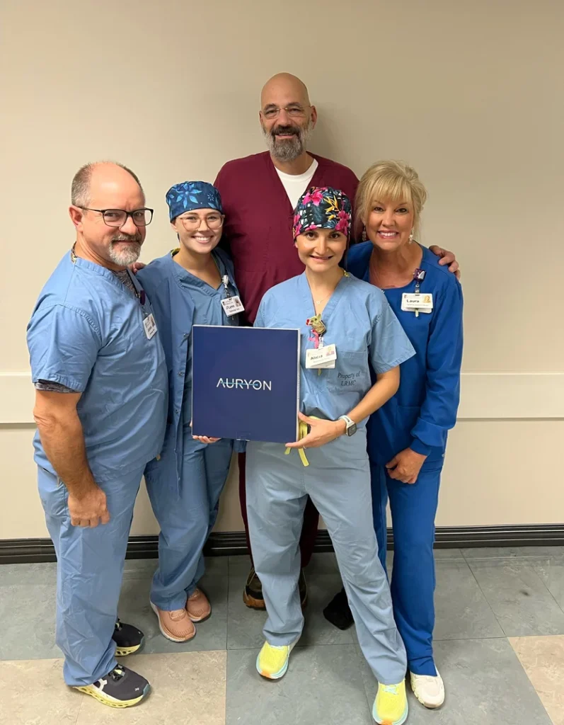 A smiling group of CIs staff members in scrubs pose with a box containing the Auryon system