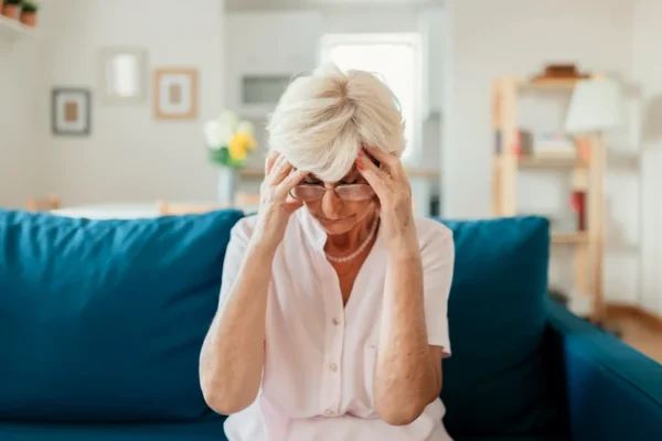 A pensive senior woman sitting on a blue sofa, holding her head in her hands, possibly in deep thought or feeling stressed about her heart health.