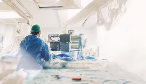 A cardiologist in scrubs and a cap focuses on a monitor displaying medical information during a procedure in a brightly lit operating room.