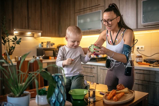 A young boy and a woman smiling in a cozy kitchen setting, as the boy excitedly reaches for a bowl that the woman, a cardiologist, is holding out to him, with ingredients on the