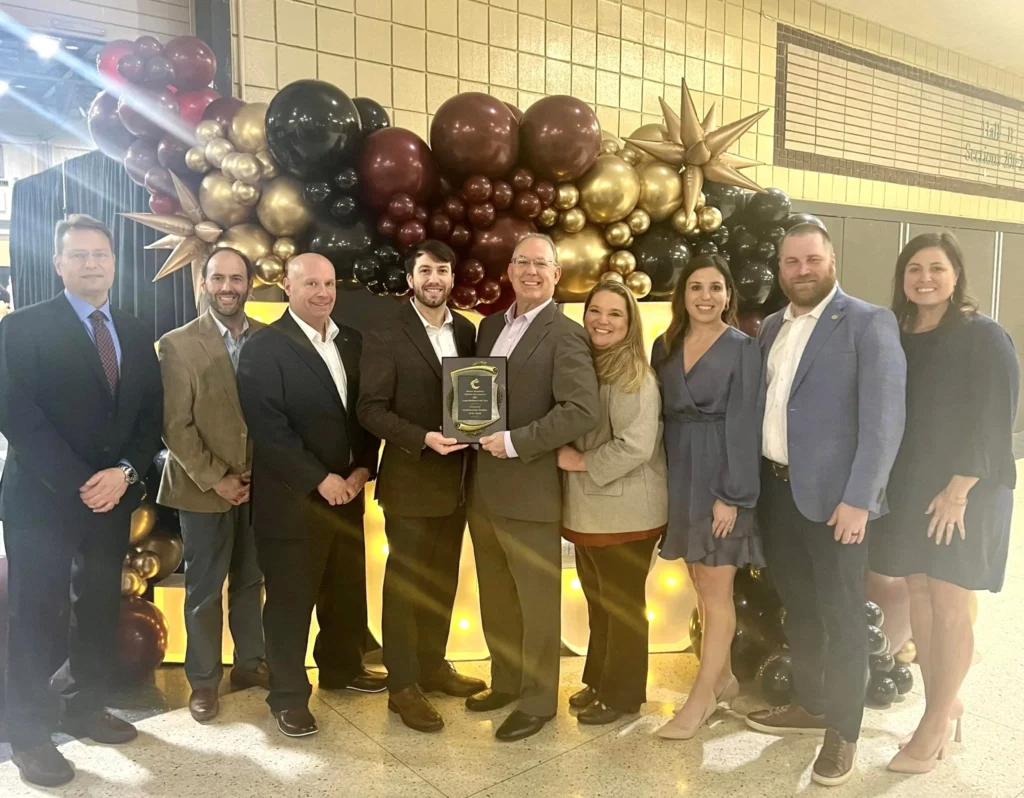 A group of professionals smiling and posing for a photo at an event, with one person holding an award plaque, standing in front of a festive balloon arrangement.