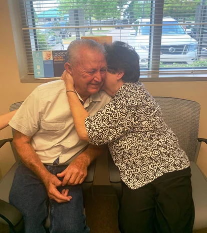 A tender moment of affection: a woman, who is also a cardiologist, kisses a man on the cheek, who appears touched by the gesture, in a room with a window view to the outside