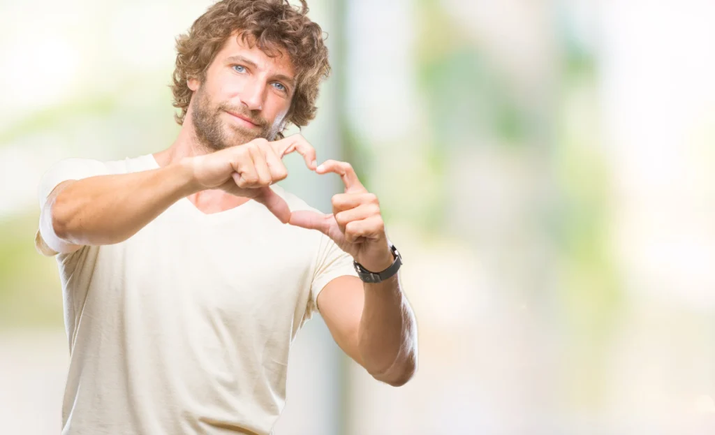 A cardiologist with curly hair forming a heart shape with his hands, smiling at the camera against a soft-focused background.