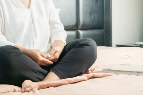 A person sitting cross-legged on a bed, practicing meditation or relaxation with a focus on their hand position for cardiology health improvement.