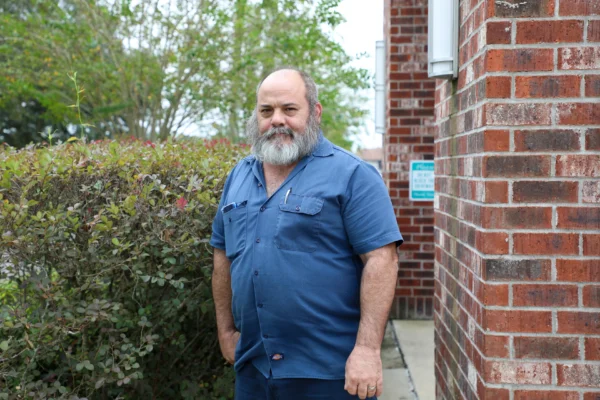 A cardiologist in a blue work uniform stands confidently outdoors beside a red brick wall, with green foliage in the background.