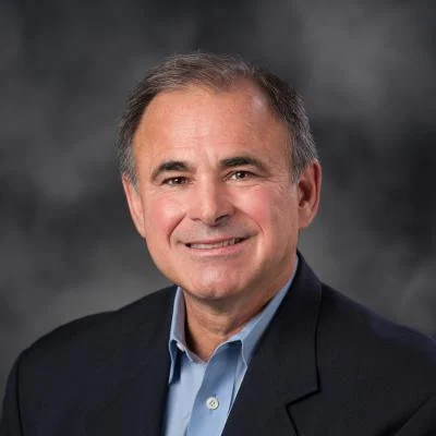 Professional headshot of a smiling heart doctor with dark hair, wearing a blue shirt and black blazer, against a mottled grey background.